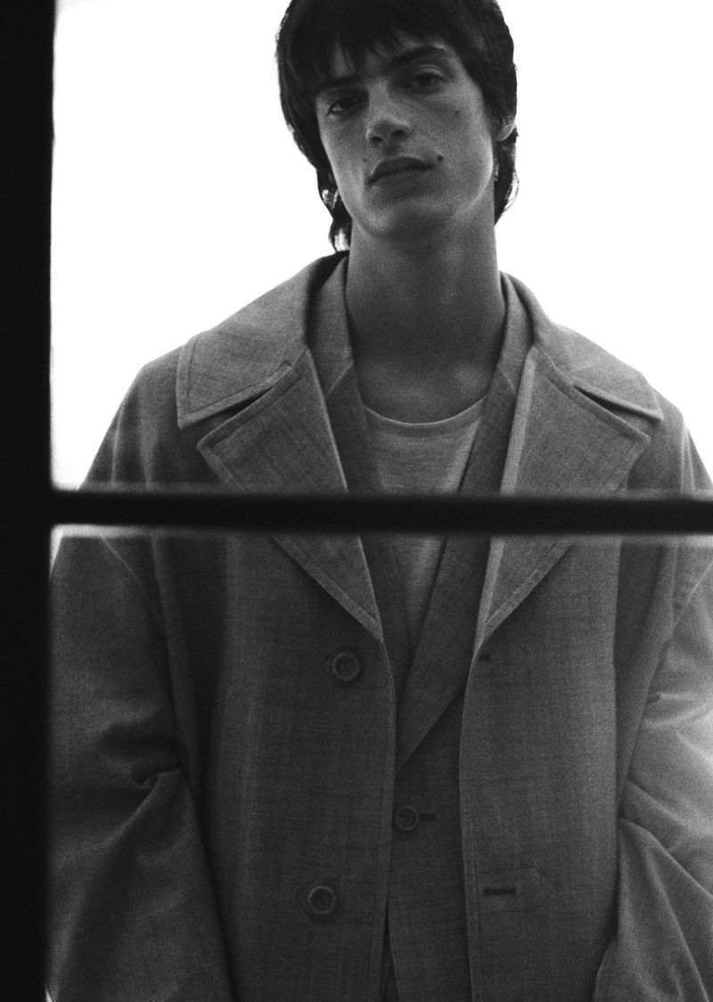 Monochromatic portrait of a young adult male in a blazer, looking at the camera indoors with natural lighting and a shallow depth of field.