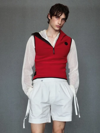 Fashion portrait of a male model wearing a sleeveless red hooded top and white shorts standing against a neutral grey background.