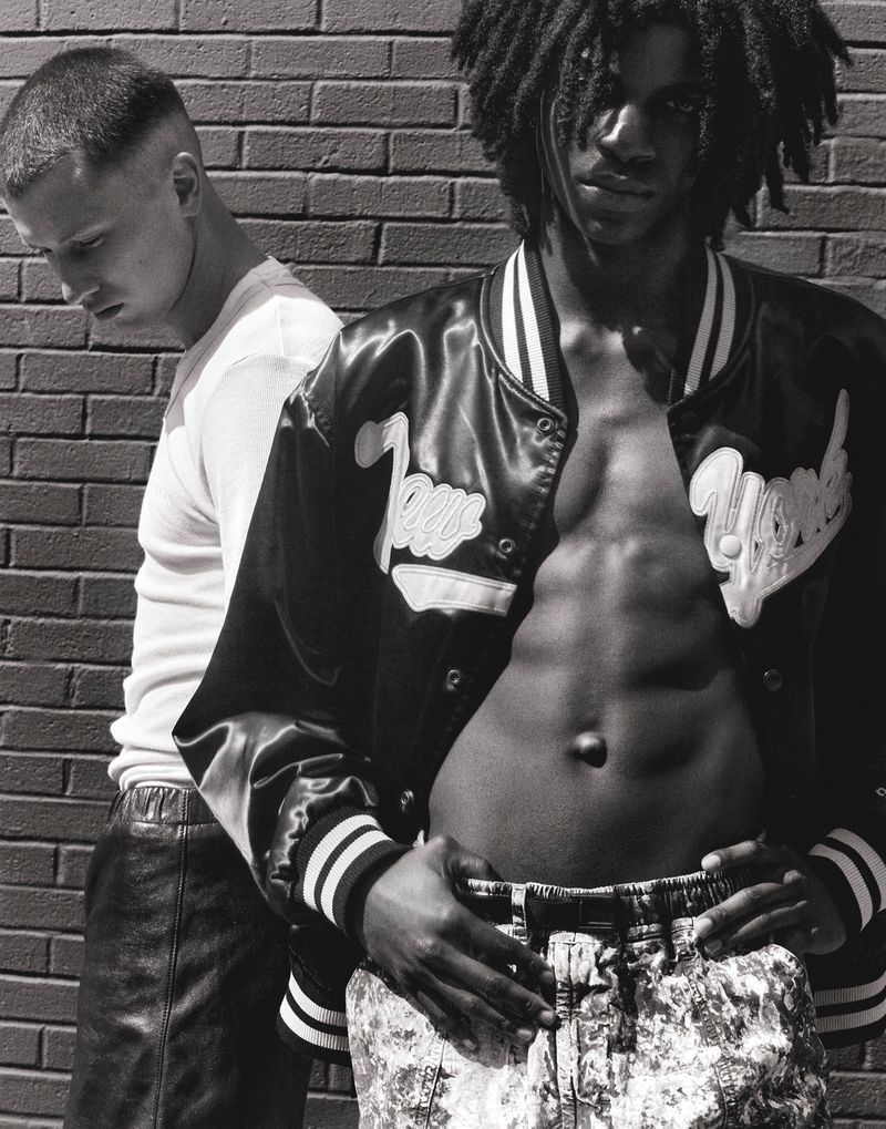 Two young men pose in front of a brick wall. One wears a letterman jacket and printed pants, while the other wears a t-shirt and leather pants.