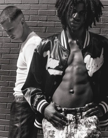 Two young men pose in front of a brick wall. One wears a letterman jacket and printed pants, while the other wears a t-shirt and leather pants.