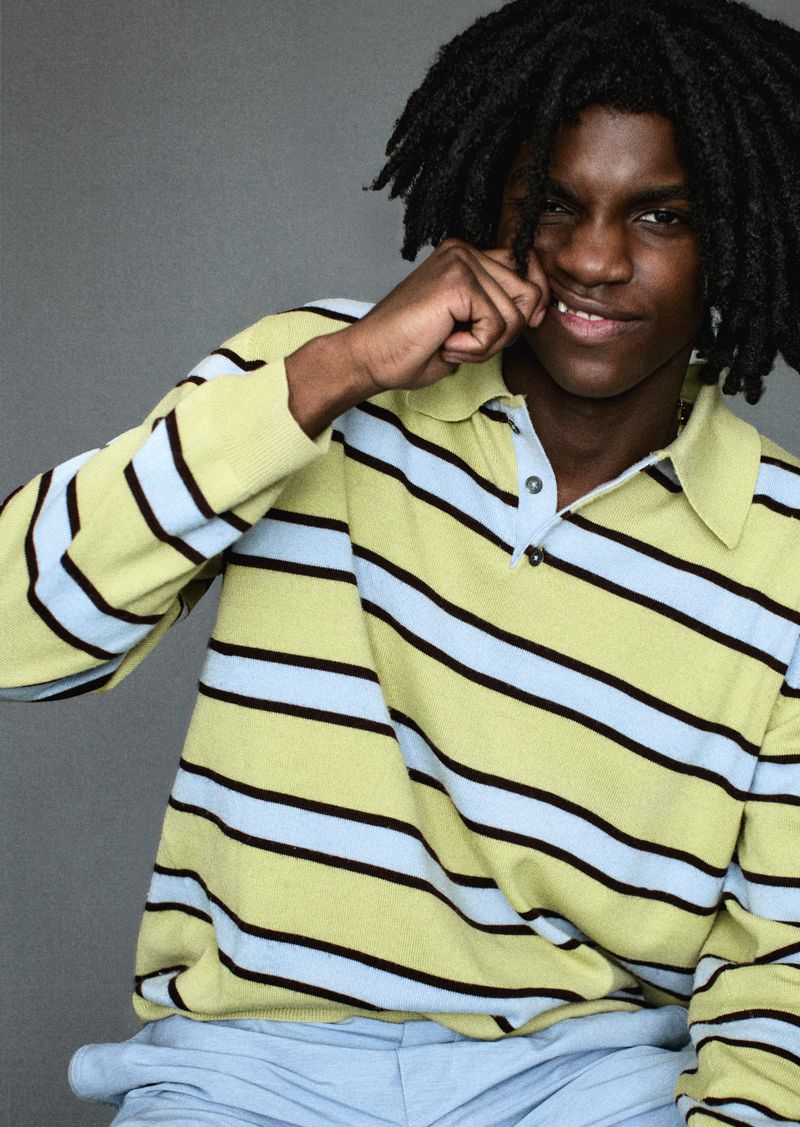 Portrait of a young adult Black man with locs, wearing a striped yellow polo shirt, playfully pinching his cheek with a smile.