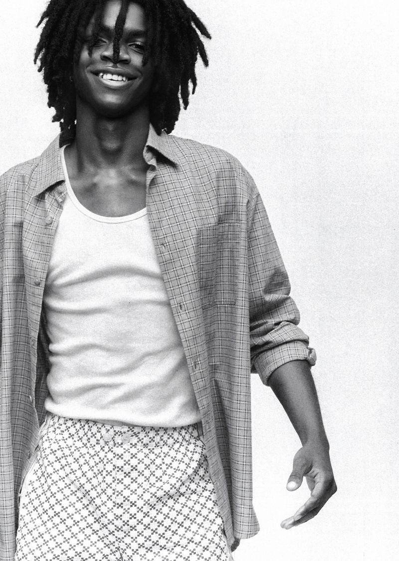 Monochrome portrait of a smiling Black man with dreadlocks wearing a white tank top and patterned pajama shorts in front of a white wall.