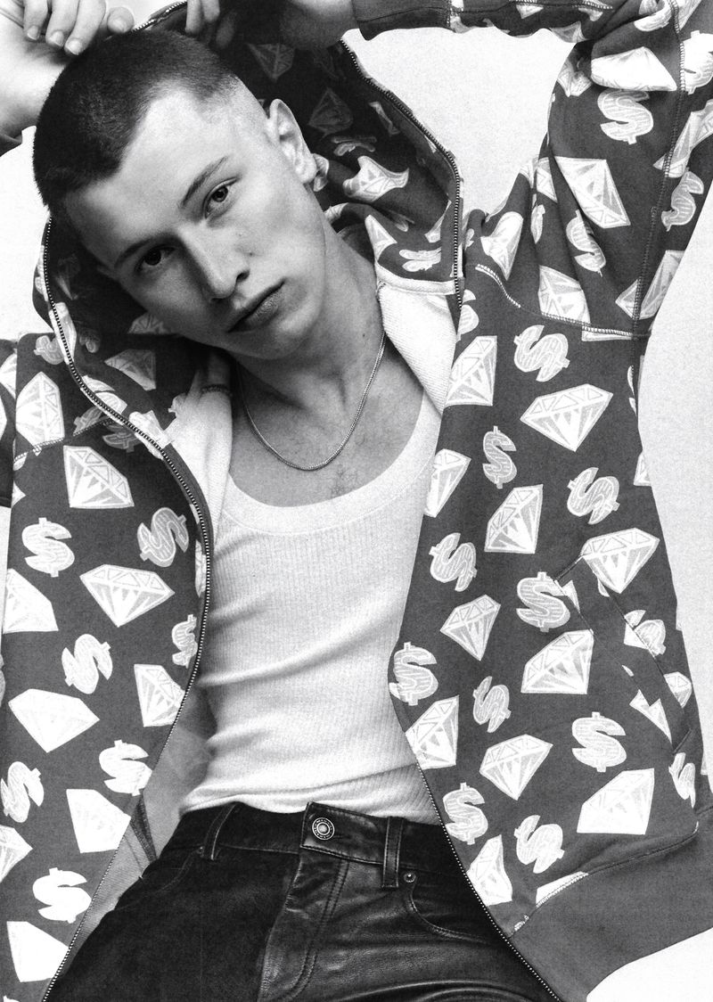 Portrait of a young adult male with short buzzcut wearing a patterned hoodie, white tank top, and dark pants against a neutral background.