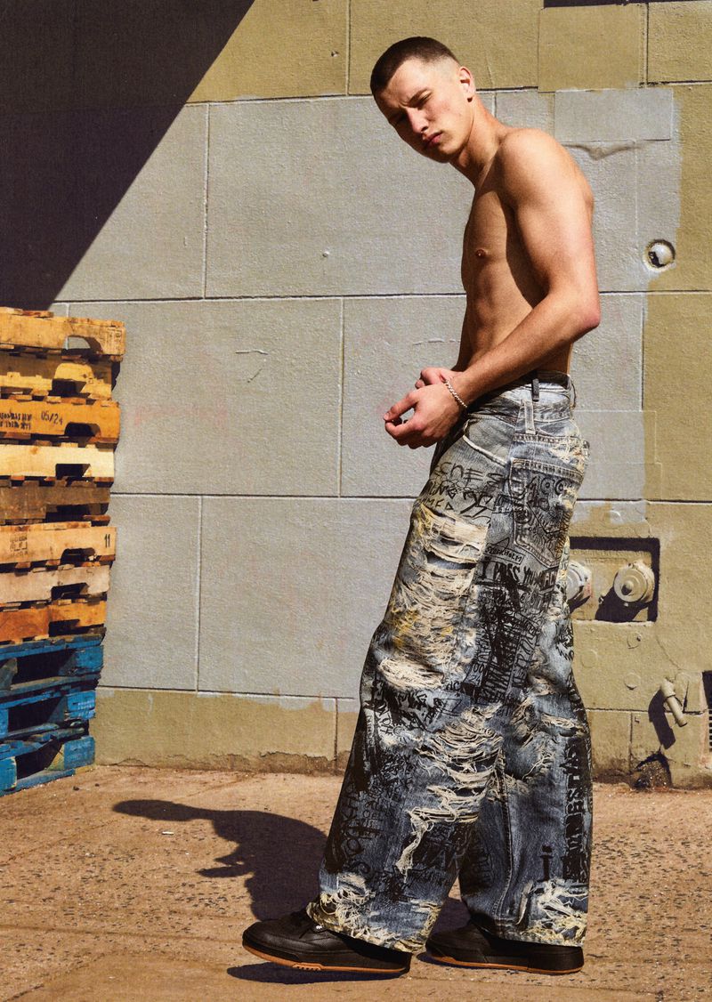A shirtless young man stands in baggy jeans outside a brick wall with wood pallets, in an outdoor fashion portrait.