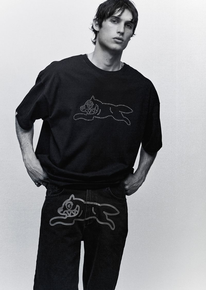 Monochrome studio shot of a young adult man modelling black T-shirt and jeans with cartoon dog design, hands in pockets, looking at camera.