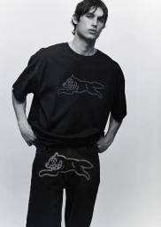 A male model wearing a black graphic t-shirt and matching jeans posing against a plain light studio background.