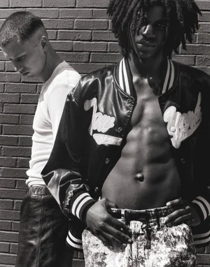 Two male models posing against a dark brick wall wearing a varsity jacket and white t-shirt in black and white