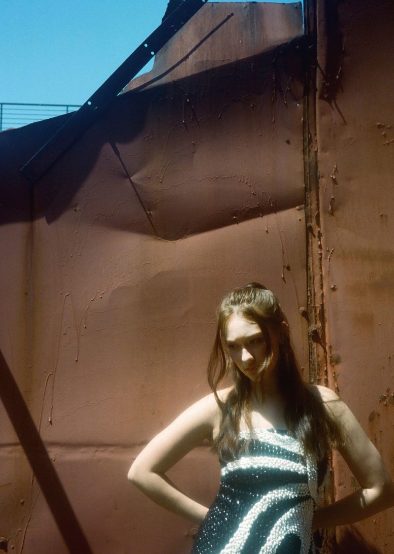 An adult female model in a striped dress poses with hands on hips against a rusty metal structure and a light blue sky.