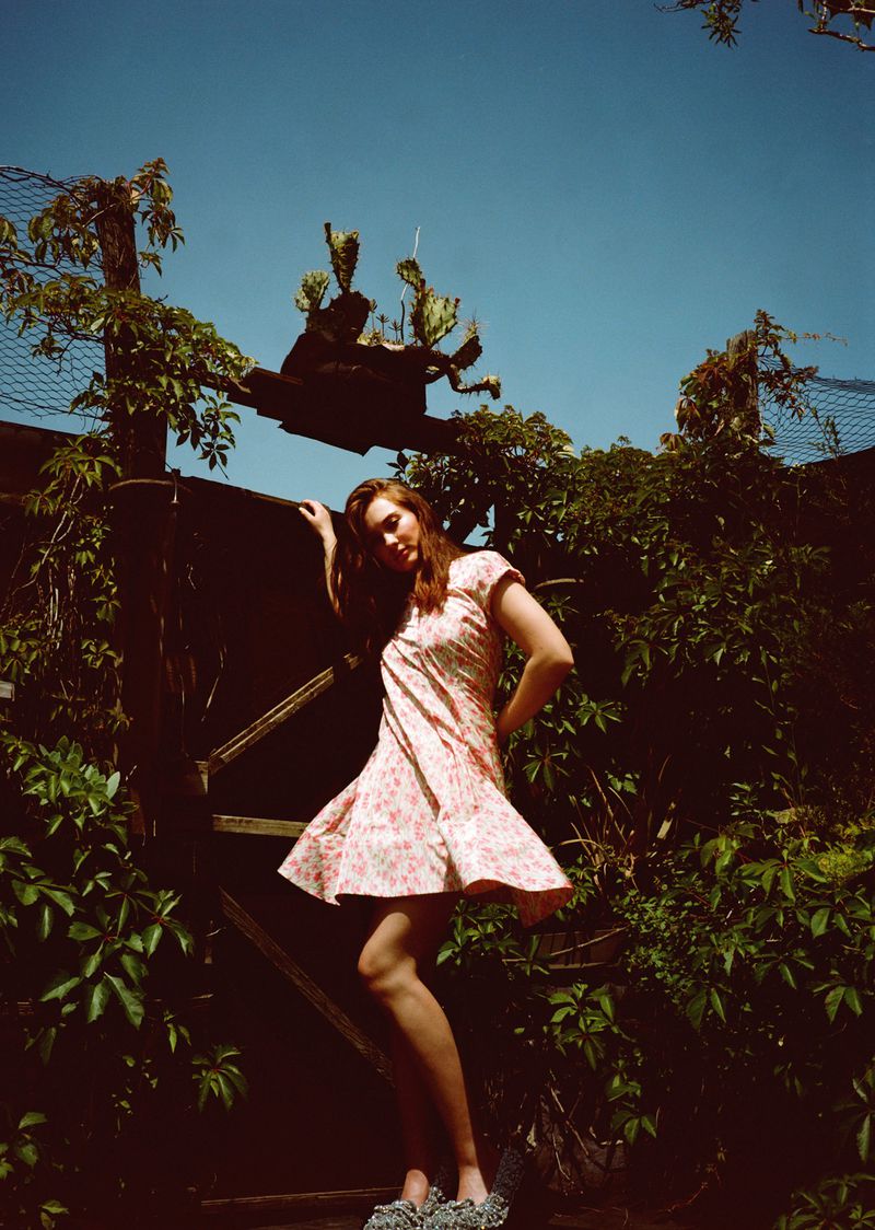 A woman in a short floral dress poses in a garden, leaning against a rustic wooden gate with a cactus and climbing vines. 