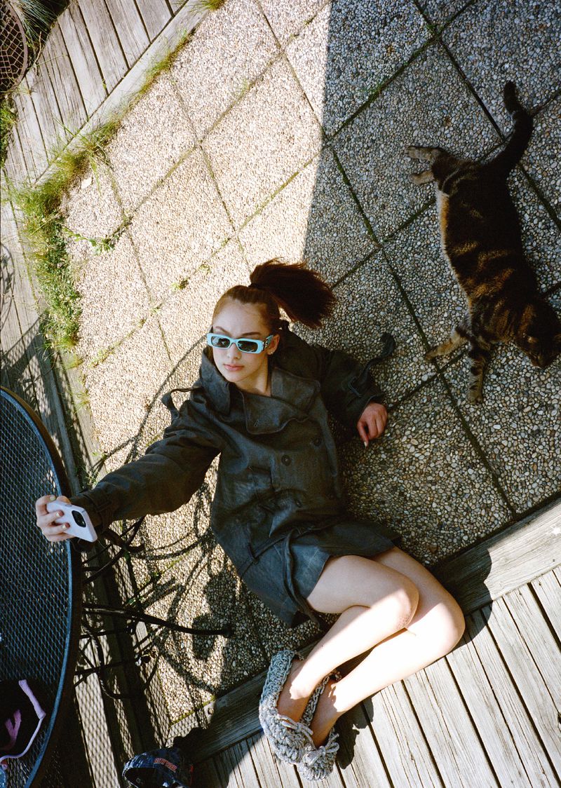 Woman takes a selfie with a cat lying on a stone terrace in the sunlight, wearing a black jacket and blue-tinted glasses.