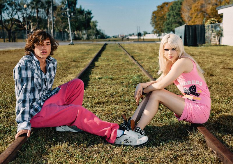 A young man and woman sit on railroad tracks in the grass, with the woman in pink and the man in plaid, posing for the camera.