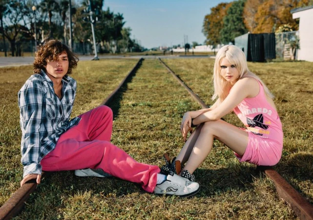 A young man and woman sit on railroad tracks in the grass, with the woman in pink and the man in plaid, posing for the camera.