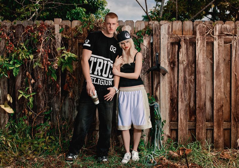 Young adult couple poses outdoors near wooden fence and lush greenery. The man wears True Religion t-shirt; the woman wears cap.