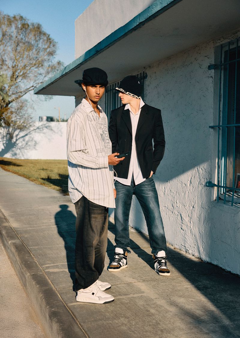 Two young men in casual street style pose near a building on a sunny day, showcasing unique hats, fashionable sneakers and denim jeans.