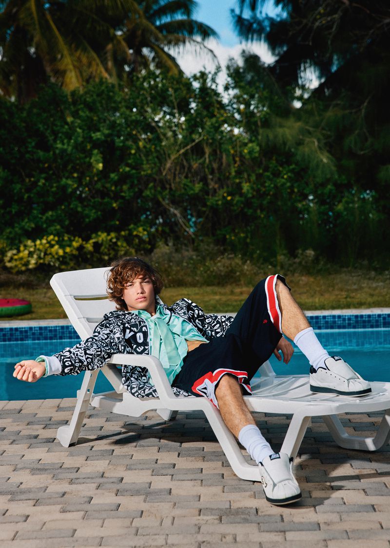 Young adult male with curly hair lounges on a white plastic chair outdoors, wearing a patterned jacket, shorts, and sneakers, with tropical plants.