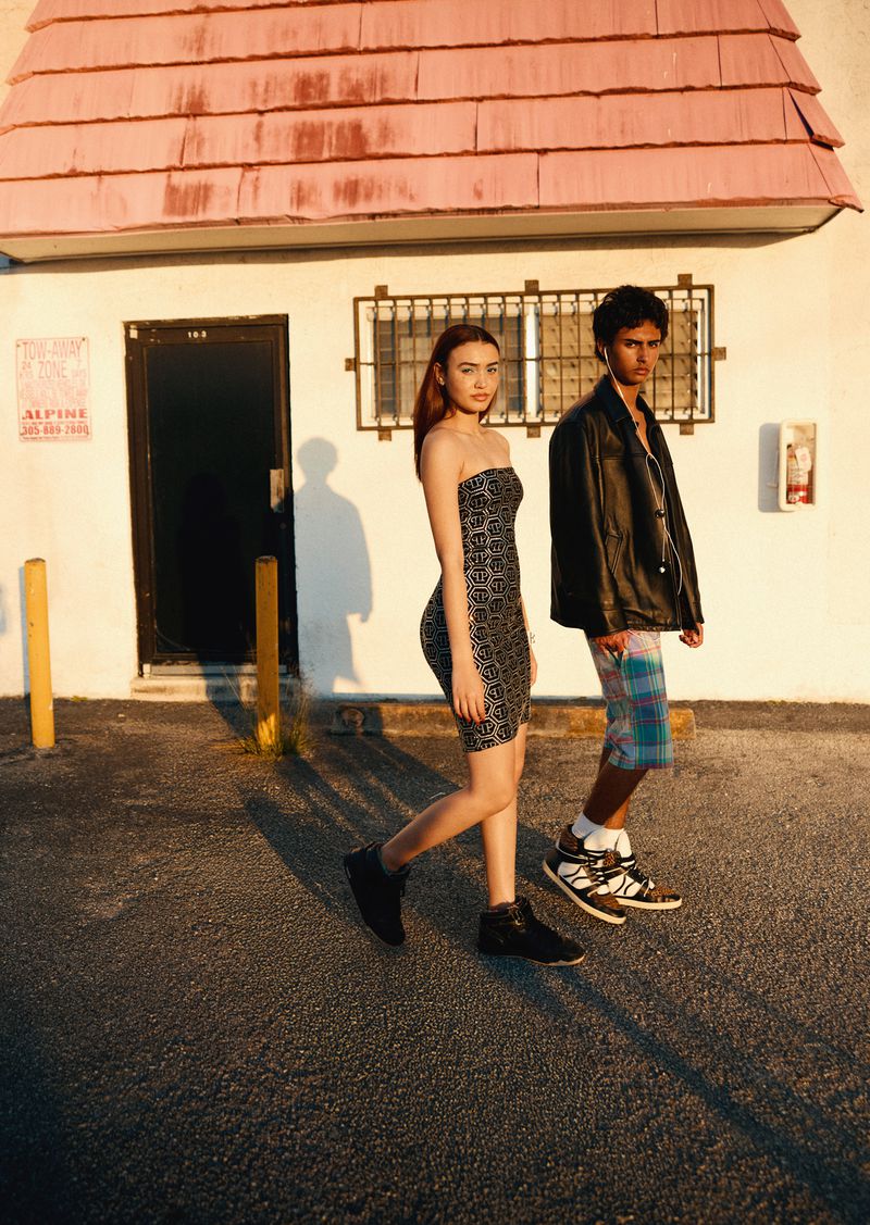 A young woman in a patterned dress and a young man in a leather jacket walk in front of a light-colored building on a sunny day.