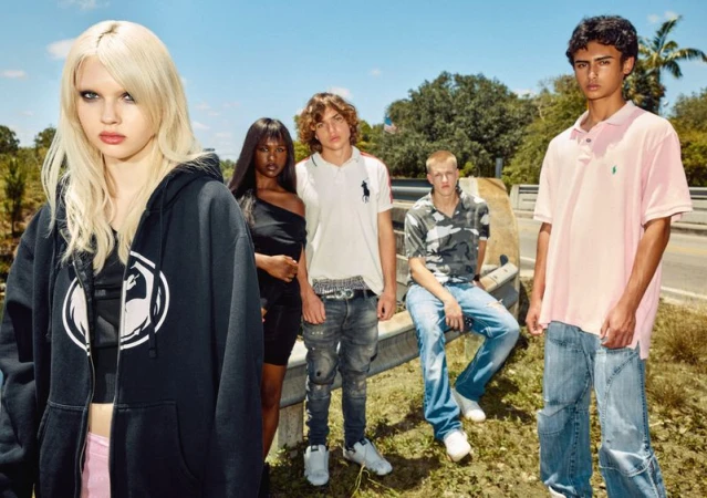 Group of five young adults outdoors wearing casual fashion apparel, posing in front of road and trees on a sunny day.