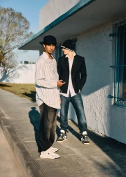 Two young men in streetwear and hats standing along an outdoor sidewalk beside a white stucco building.