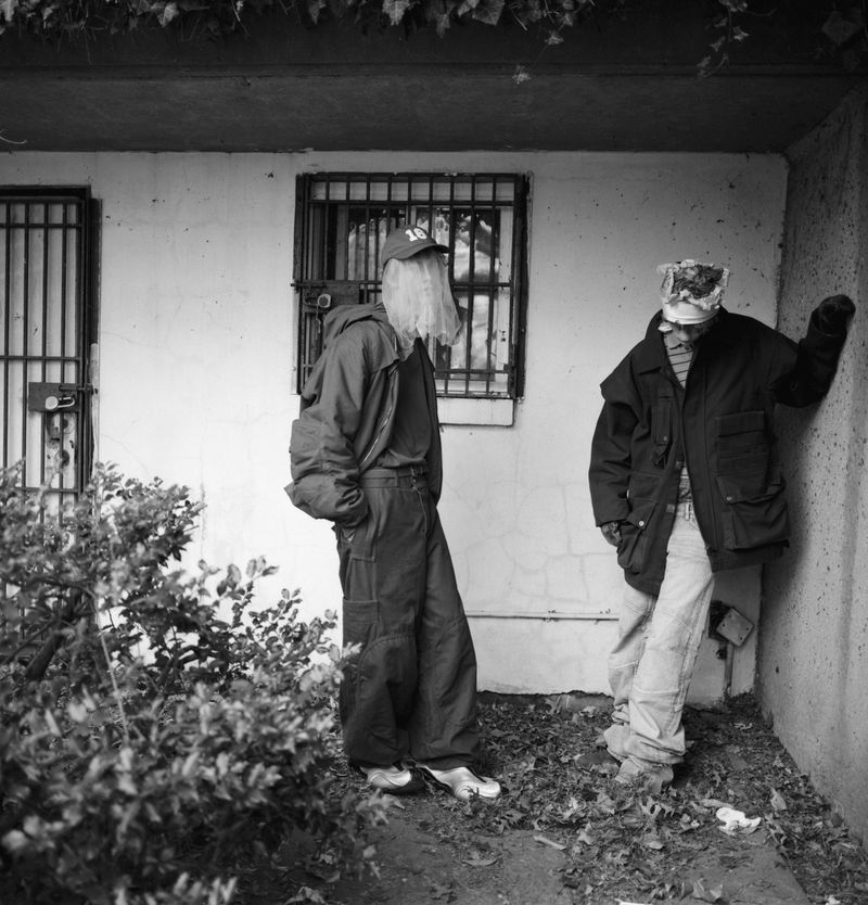 Black and white shot of two individuals in casual outerwear standing in an alleyway beside a wall with barred windows and ivy.
