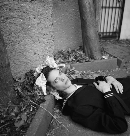 A young woman lies in a sailor-style jacket next to a concrete barrier outdoors, decorated with light-colored bows in her hair.