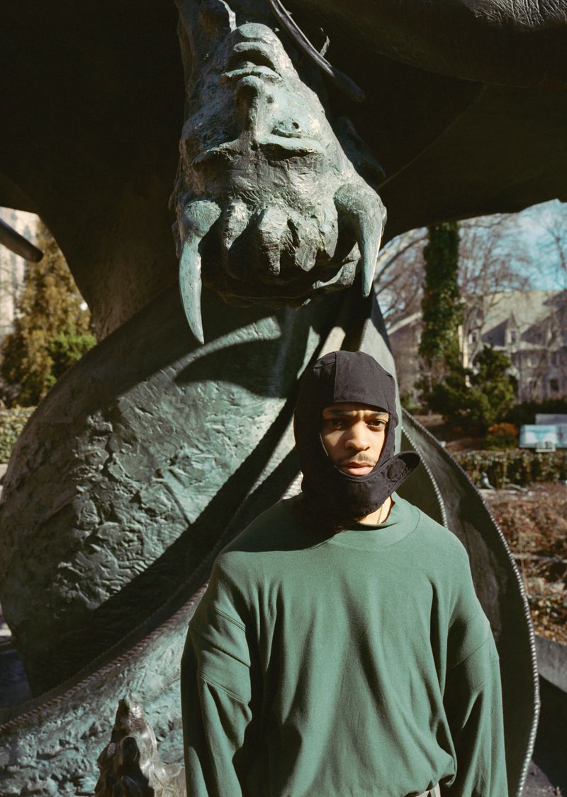 Young adult poses outdoors in a dark green sweater and head covering against a backdrop of a bronze dragon statue and bare trees.