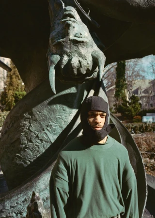 Young adult poses outdoors in a dark green sweater and head covering against a backdrop of a bronze dragon statue and bare trees.