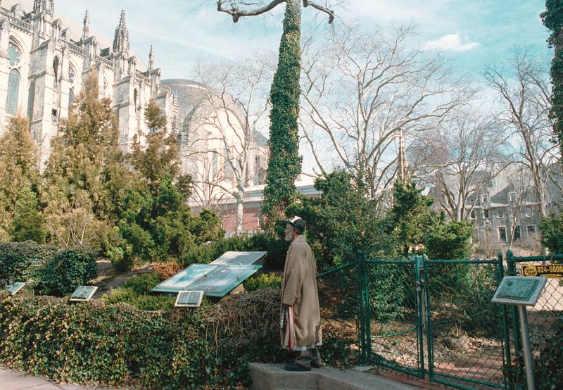 Adult male wearing a tan coat and hat stands next to a garden of green plants and a metal fence with a cathedral visible in the background.