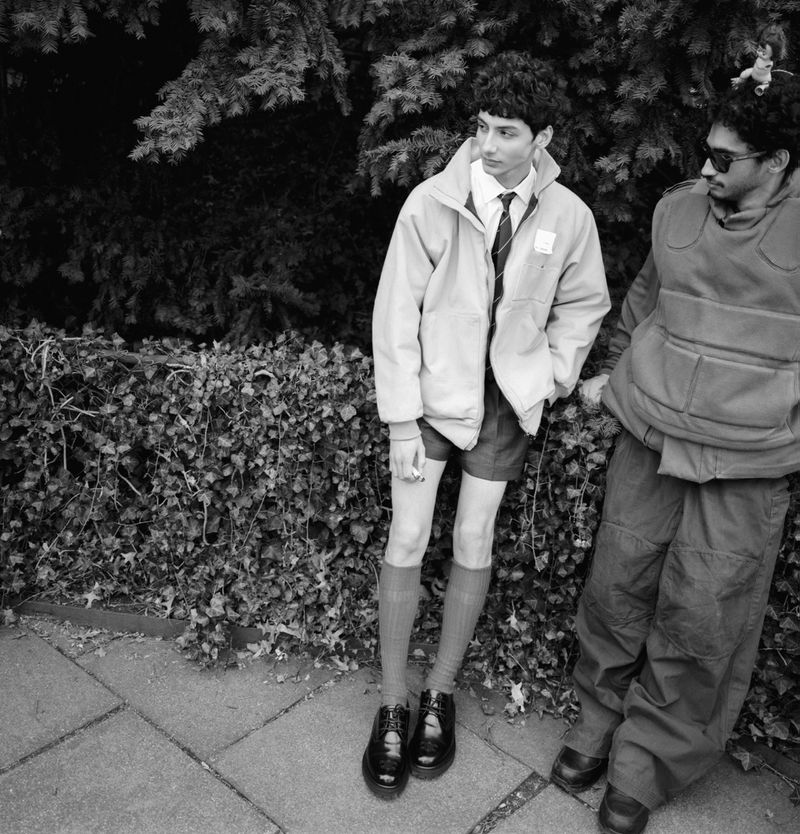 Two young men stand outdoors against green foliage. One wears a light jacket, shorts, and high socks. The other wears a vest and pants.