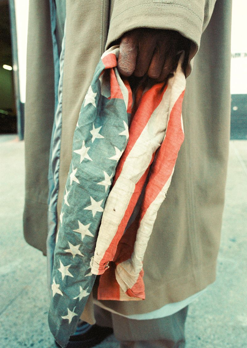 A man's hand holds a tattered American flag against a neutral city background, with focus on the texture and colors of the flag.