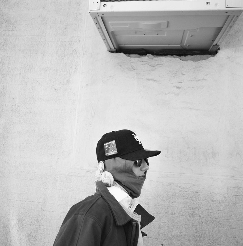 Monochrome portrait of a person in sunglasses, a cap, and a balaclava, against a textured wall with a metal box overhead.