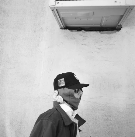 Monochrome portrait of a person in sunglasses, a cap, and a balaclava, against a textured wall with a metal box overhead.