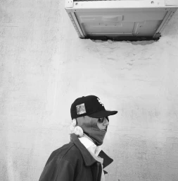 Monochrome profile portrait of a model wearing a black baseball cap, sunglasses, balaclava, and jacket against an exterior wall.