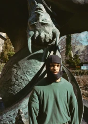 Model Daniel Abramson wearing a green long-sleeve shirt and black balaclava posed in front of a bronze horned statue outdoors.