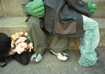 Model wearing a decorative headpiece lying on concrete next to seated figures wearing green monster hands for HERO Summer Zine.