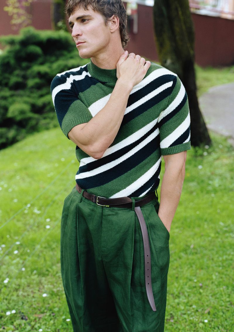 Man in striped sweater and green pants posing outdoors in a park-like setting, looking away from the camera.