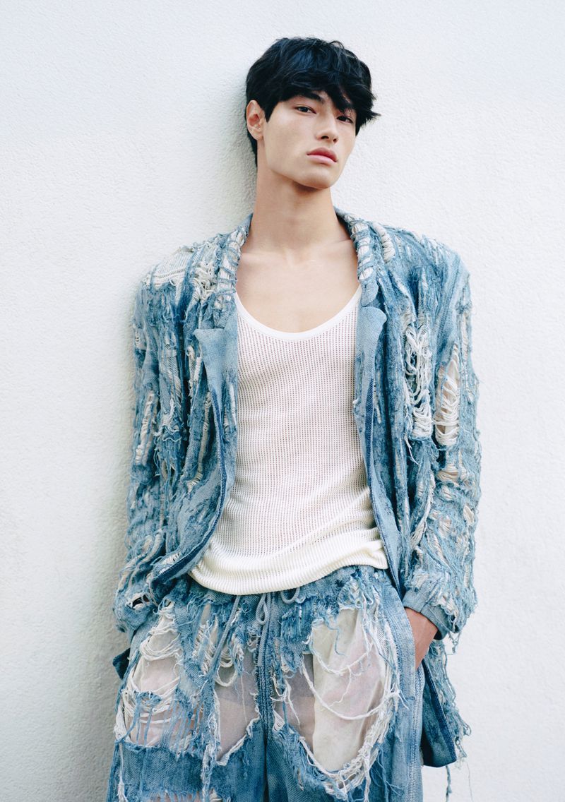 Male fashion model wearing a shredded denim blazer and shorts set and a white tank top poses against a white wall indoors.