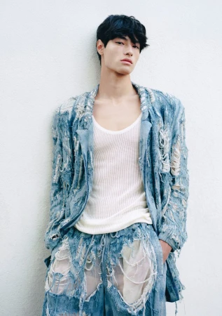 Male fashion model wearing a shredded denim blazer and shorts set and a white tank top poses against a white wall indoors.
