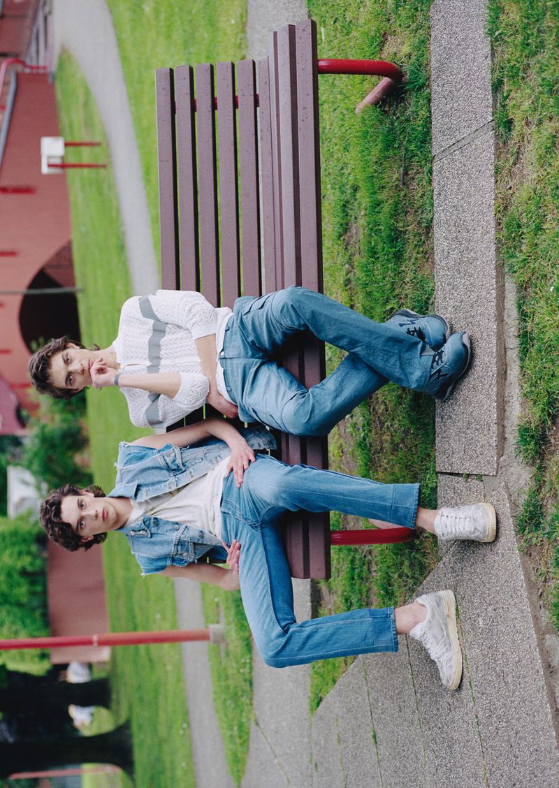 Two young men in casual denim and knitwear are posing on a wooden park bench in an outdoor green space with trees and a red brick structure.