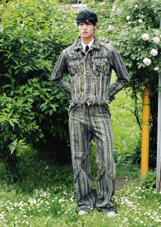 A young man poses outdoors wearing a distressed plaid denim jacket with matching pants, standing on a grassy area surrounded by greenery.
