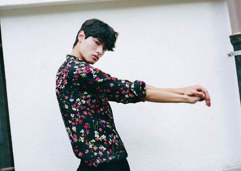 Young adult male posing in a floral print shirt in front of a white wall, creating a fashionable and casual outdoor portrait.
