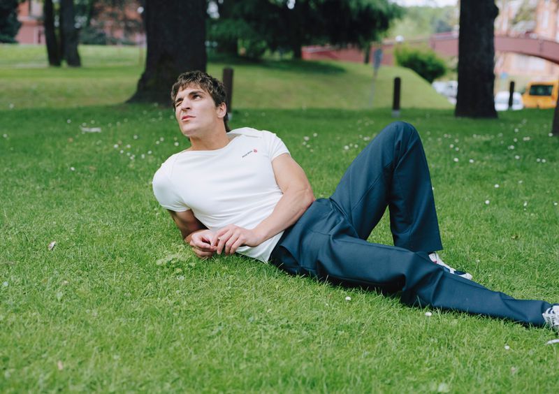 Adult male lying on grass in an outdoor park setting wearing a white short-sleeve shirt and blue pants, looking upwards.