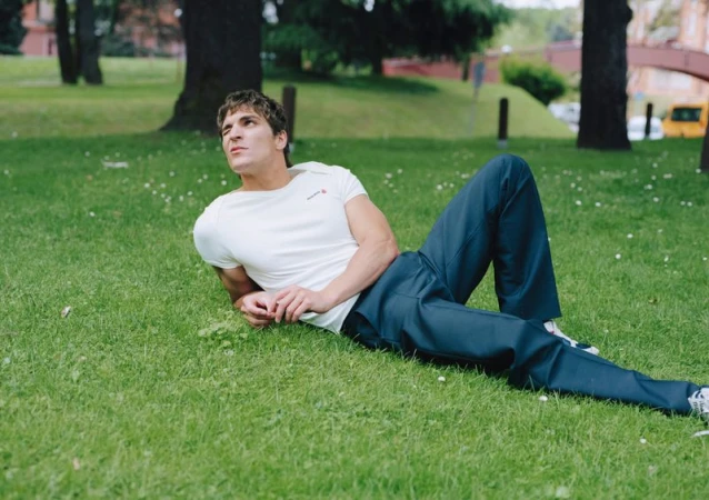 Adult male lying on grass in an outdoor park setting wearing a white short-sleeve shirt and blue pants, looking upwards.