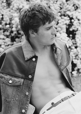 Black and white portrait of a male model with an open denim jacket in front of a flowering bush for fashion or lifestyle content.