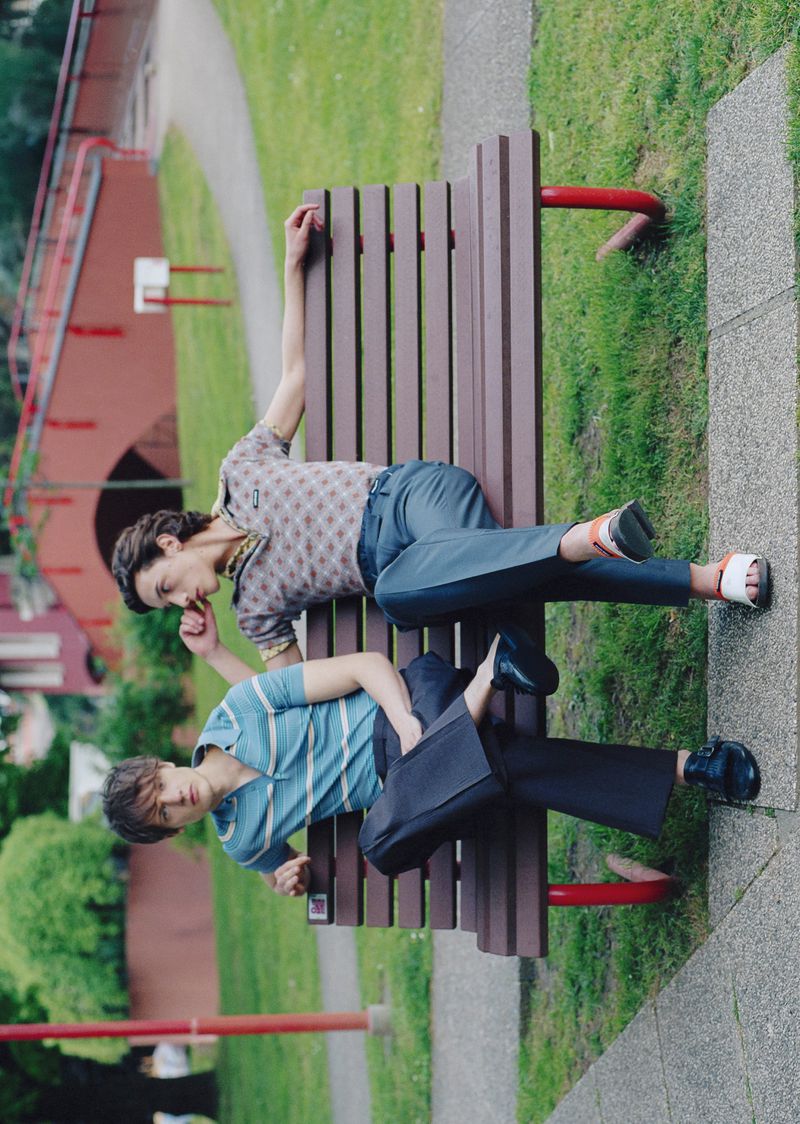 Two people relax on a park bench in casual clothes. One person leans back while the other sits upright in a green urban park setting.
