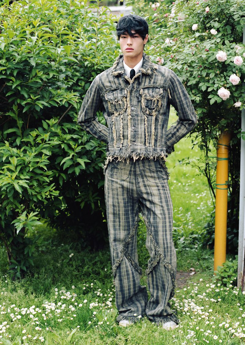 East Asian male model wearing a distressed plaid jacket and matching trousers standing in a lush garden.