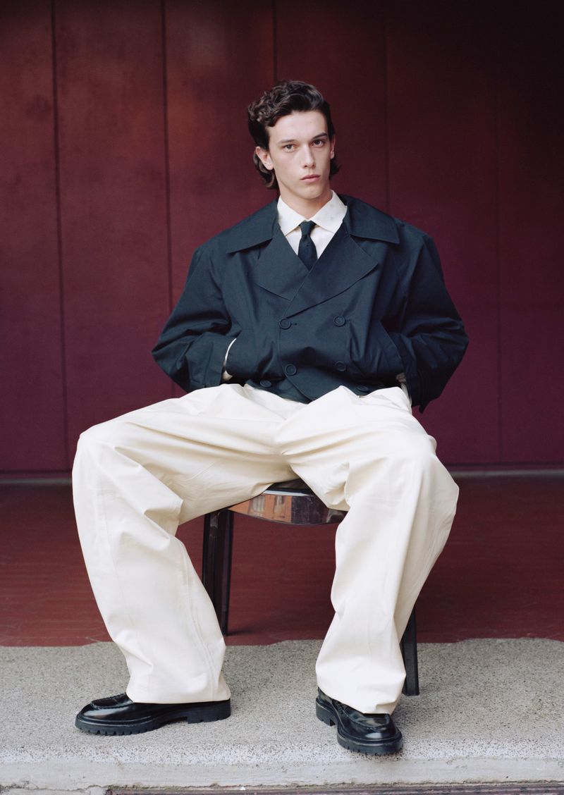 Seated male model wearing a dark cropped jacket, black tie, and wide-leg cream trousers against a burgundy paneled wall.