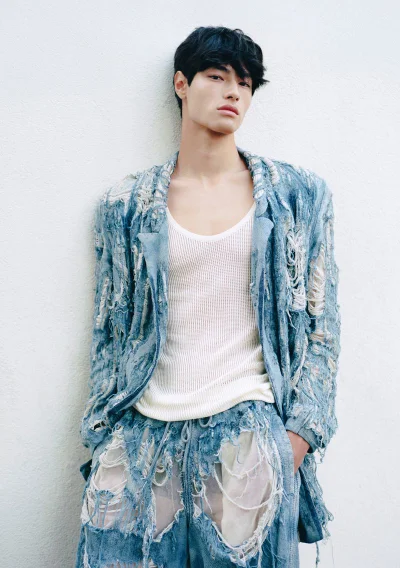Model wearing a heavily distressed light blue denim jacket and ripped trousers with a white ribbed tank against a textured wall.