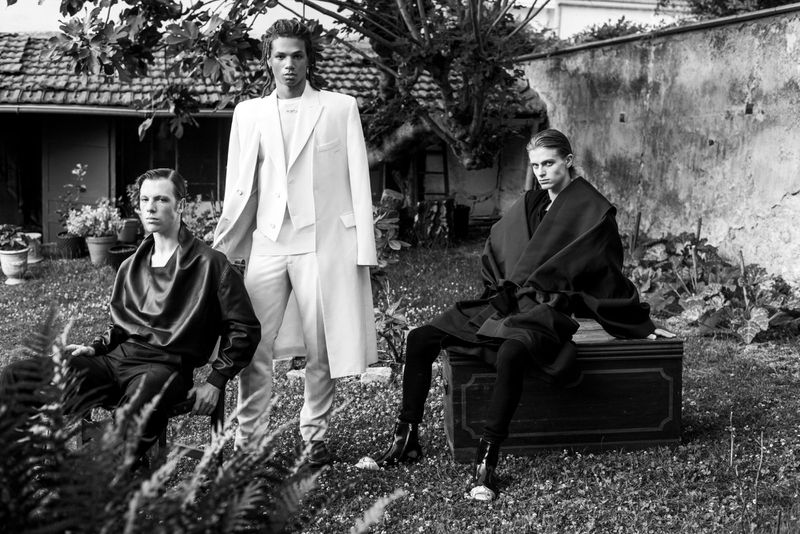 Three fashion models in black and white stand outdoors in high-fashion clothing against a backdrop of plants and a rustic building.