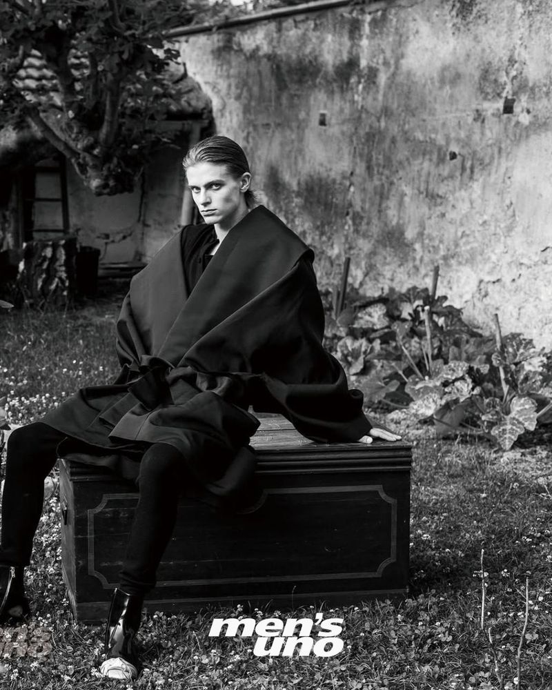 Monochrome fashion shot: Man in oversized coat poses on trunk outdoors. Black and white image of outdoor fashion shoot.