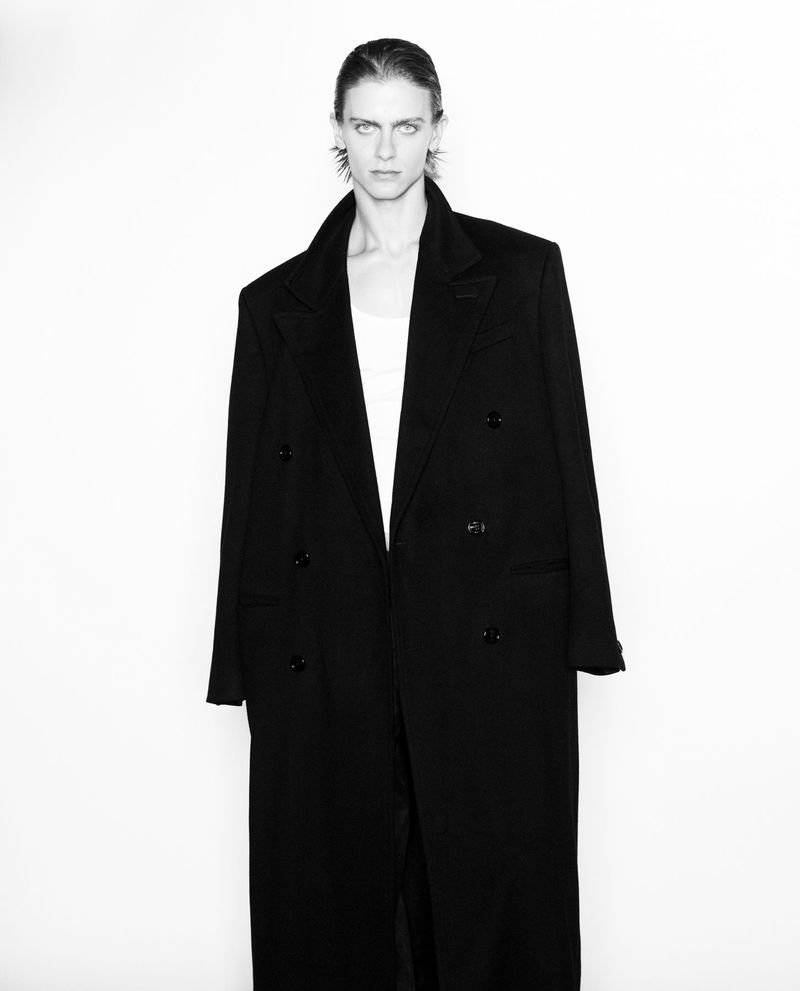 Black and white studio portrait of a slender adult with neutral expression, wearing a long, dark overcoat and a white top.
